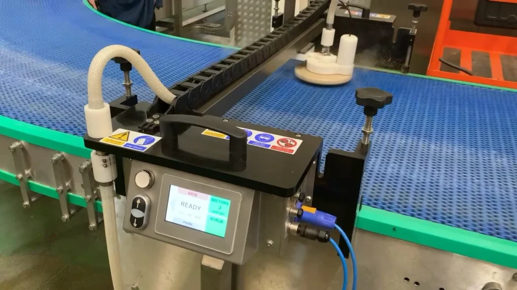 Automated modular conveyor belt cleaning system using dry steam, rotating cleaning head and vacuum extraction on a blue modular plastic conveyor belt.